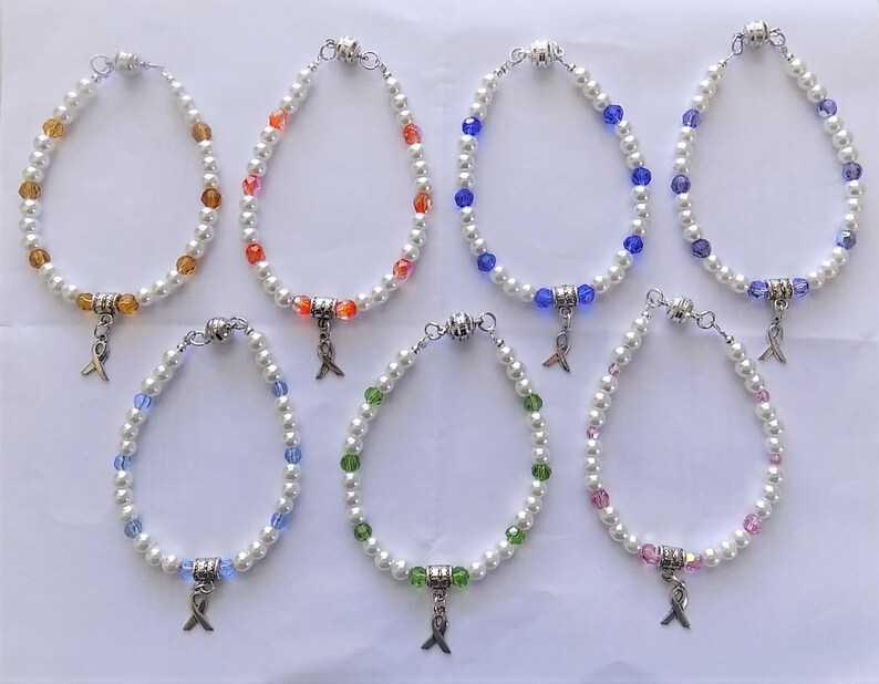 May include: Eight pearl bracelets with different colored beads and a silver charm in the shape of a ribbon. The bracelets are arranged in two rows of four.