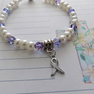 May include: A white pearl and purple crystal bracelet with a silver ribbon charm. The bracelet is on a white lined paper background with a drawing of a cross with flowers.