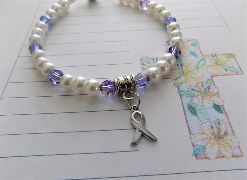 May include: A white pearl and purple crystal bracelet with a silver ribbon charm. The bracelet is on a white lined paper background with a drawing of a cross with flowers.
