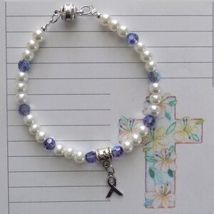 May include: A white pearl bracelet with purple glass beads and a silver charm with a purple ribbon. The bracelet has a magnetic clasp.