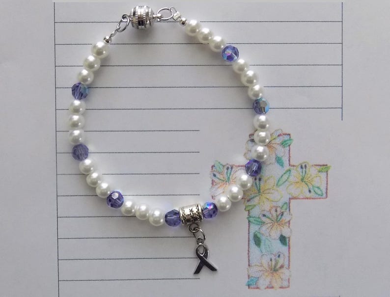 May include: A white pearl bracelet with purple glass beads and a silver charm with a purple ribbon. The bracelet has a magnetic clasp.