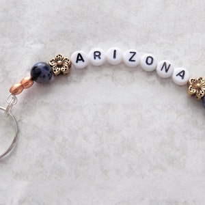 May include: A silver key chain with a blue and white stone bead, gold flower charm, and white letter beads spelling "ARIZONA".