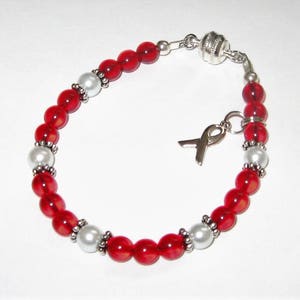 May include: Red and white beaded bracelet with a silver charm in the shape of a ribbon. The bracelet is made with small red glass beads and white pearl beads.