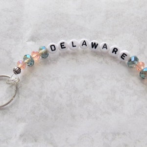 May include: A white beaded keychain with the word "DELAWARE" spelled out in black letters. The beads are interspersed with clear and pink crystal beads.