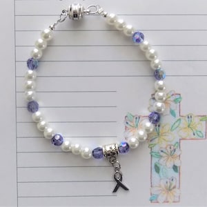 May include: A white pearl bracelet with purple crystal beads and a silver charm with a purple ribbon. The bracelet has a magnetic clasp.
