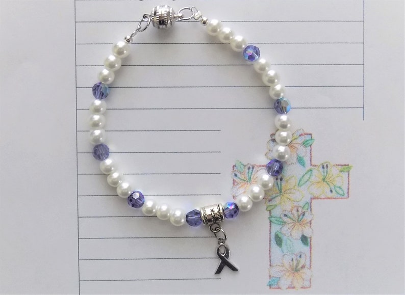 May include: A white pearl bracelet with purple crystal beads and a silver charm with a purple ribbon. The bracelet has a magnetic clasp.