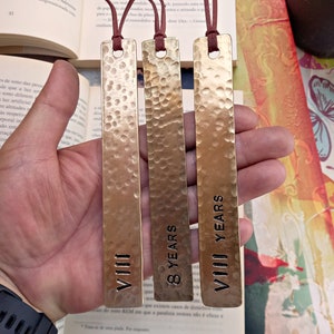 Può includere: Tre segnalibri in metallo color oro con scritte nere. I segnalibri sono martellati e hanno una superficie testurizzata. Il testo sui segnalibri recita "VIII", "8 YEARS" e "VIII YEARS".