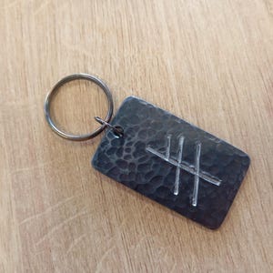 Könnte beinhalten: Ein silberfarbener Schlüsselanhänger aus Metall mit gehämmerter Textur. Der Schlüsselanhänger zeigt ein Symbol eines Kreuzes mit einer vertikalen Linie in der Mitte.