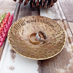 Może przedstawiać: Mosiężna podstawka na pierścionki o młotkowanej fakturze. Podstawka ma napis "IIII Years & Counting..." i małe serce. Dwa pierścionki leżą na podstawce.