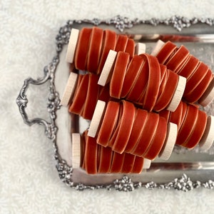 May include: Spools of rust-colored velvet ribbon are displayed on a decorative silver tray. The ribbon is tightly wound around small wooden spools. The tray has ornate handles and edges, set against a white, patterned background.