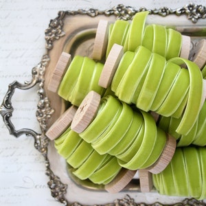 May include: A close-up of spools of lime green velvet ribbon on a silver tray. The ribbon is wound around wooden spools. EAB Designs Pemberley Collection is written on the background.