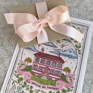 K&ouml;nnte beinhalten: Eine braune Geschenkbox mit einem rosa Satinband, das zu einer Schleife gebunden ist. Die Box liegt auf einer Karte mit einer Illustration eines rosa Hauses und dem Text "La maison du bonheur".