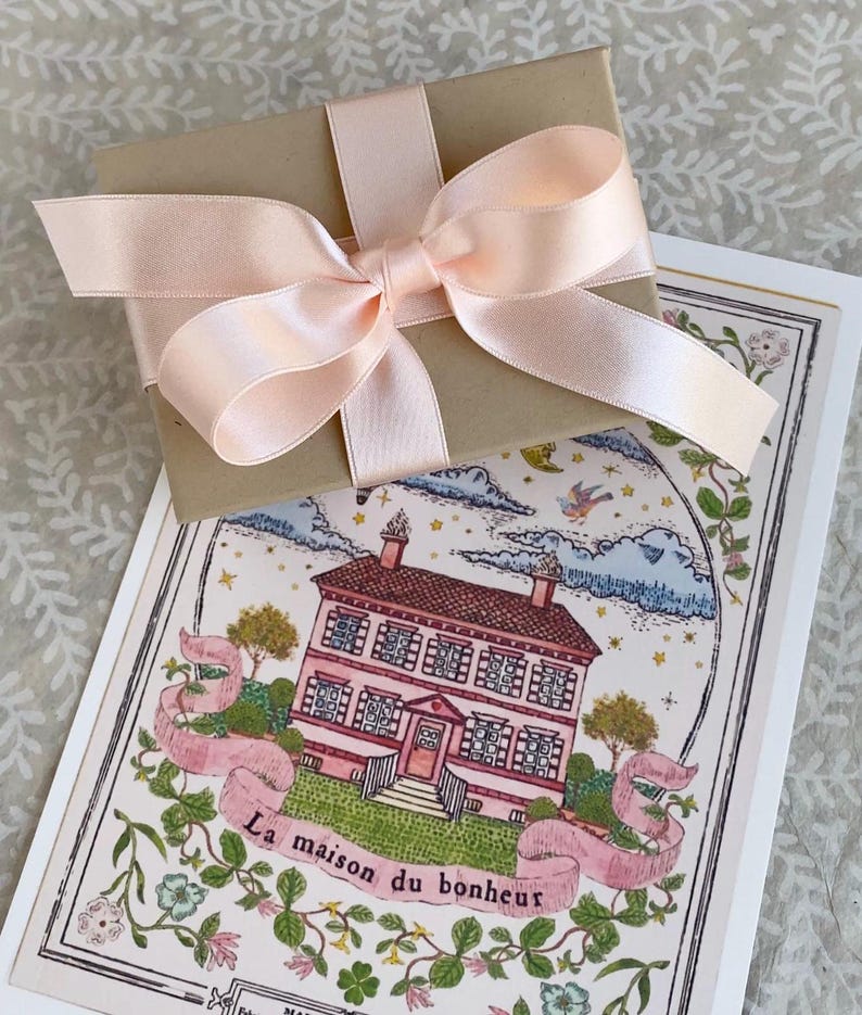 K&ouml;nnte beinhalten: Eine braune Geschenkbox mit einem rosa Satinband, das zu einer Schleife gebunden ist. Die Box liegt auf einer Karte mit einer Illustration eines rosa Hauses und dem Text "La maison du bonheur".