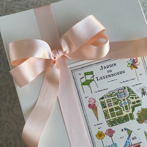 K&ouml;nnte beinhalten: Eine wei&szlig;e Geschenkbox mit einem rosa Satinband, das zu einer Schleife gebunden ist. Die Box hat eine Karte, die mit einer bunten Illustration des Jardin du Luxembourg in Paris, Frankreich, versehen ist. Die Karte enth&auml;lt Text in Franz&ouml;sisch, darunter den Namen des Parks, "Jardin du Luxembourg", und die Nummer "25".
