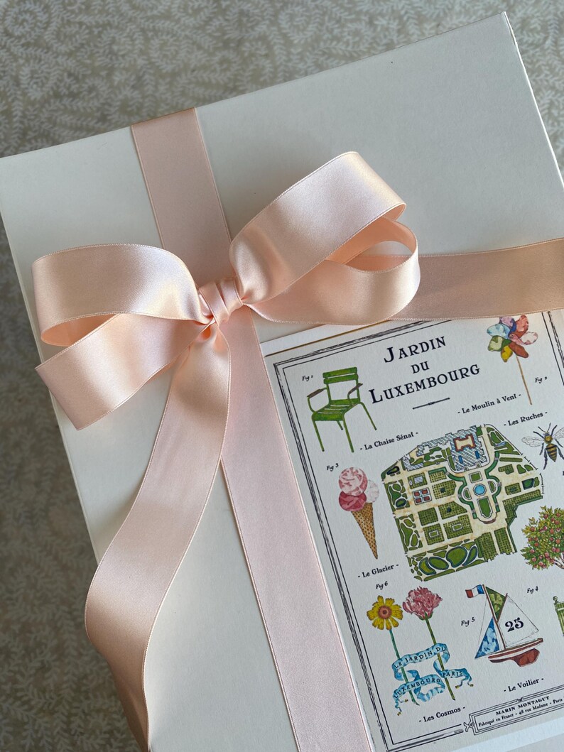 K&ouml;nnte beinhalten: Eine wei&szlig;e Geschenkbox mit einem rosa Satinband, das zu einer Schleife gebunden ist. Die Box hat eine Karte, die mit einer bunten Illustration des Jardin du Luxembourg in Paris, Frankreich, versehen ist. Die Karte enth&auml;lt Text in Franz&ouml;sisch, darunter den Namen des Parks, "Jardin du Luxembourg", und die Nummer "25".
