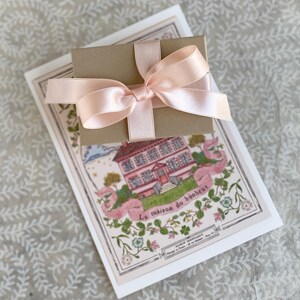 K&ouml;nnte beinhalten: Eine kleine braune Geschenkbox, die mit einem rosa Satinband gebunden ist. Die Box liegt auf einer wei&szlig;en Karte mit einer Illustration eines rosa Hauses und dem Text "La maison du bonheur".
