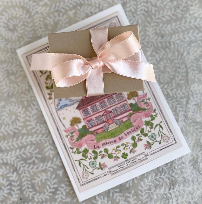 K&ouml;nnte beinhalten: Eine kleine braune Geschenkbox, die mit einem rosa Satinband gebunden ist. Die Box liegt auf einer wei&szlig;en Karte mit einer Illustration eines rosa Hauses und dem Text "La maison du bonheur".