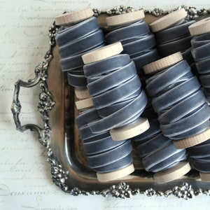 May include: A close-up of several spools of gray velvet ribbon, each wound around a wooden spool. The spools are arranged in a silver tray.