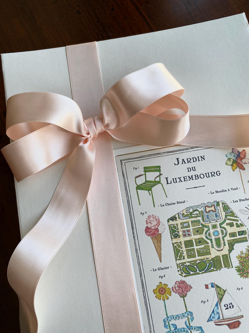 K&ouml;nnte beinhalten: Eine wei&szlig;e Geschenkbox mit einem rosa Satinband, das zu einer Schleife gebunden ist. Die Box hat eine Karte mit einer Karte des Jardin du Luxembourg in Paris, Frankreich, befestigt. Die Karte zeigt verschiedene Sehensw&uuml;rdigkeiten und Merkmale des Parks, darunter den Eisstand, den Brunnen und den Stuhl.