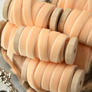 May include: Peach-colored velvet ribbon spools wrapped around wooden bobbins. The ribbon is arranged in a stack, with the bobbins visible.
