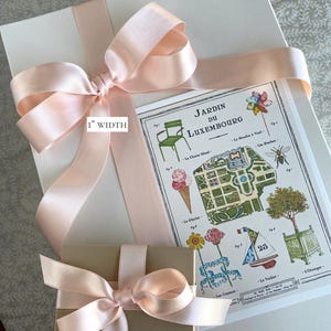 K&ouml;nnte beinhalten: Ein hellrosa Satinband, das zu einer Schleife auf einem wei&szlig;en Geschenkpaket gebunden ist. Ein kleinerer brauner Geschenkkarton mit einem hellrosa Satinband, das zu einer Schleife gebunden ist, befindet sich vor dem gr&ouml;&szlig;eren Karton. Der kleinere Karton ist mit "5/8" WIDTH" beschriftet. Der gr&ouml;&szlig;ere Karton ist mit "1" WIDTH" beschriftet. Im Hintergrund ist eine Karte des Jardin du Luxembourg in Paris zu sehen.