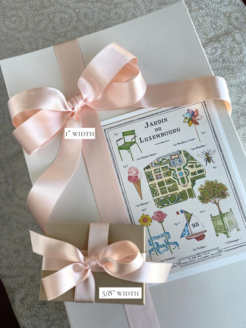 K&ouml;nnte beinhalten: Ein hellrosa Satinband, das zu einer Schleife auf einem wei&szlig;en Geschenkpaket gebunden ist. Ein kleinerer brauner Geschenkkarton mit einem hellrosa Satinband, das zu einer Schleife gebunden ist, befindet sich vor dem gr&ouml;&szlig;eren Karton. Der kleinere Karton ist mit "5/8" WIDTH" beschriftet. Der gr&ouml;&szlig;ere Karton ist mit "1" WIDTH" beschriftet. Im Hintergrund ist eine Karte des Jardin du Luxembourg in Paris zu sehen.