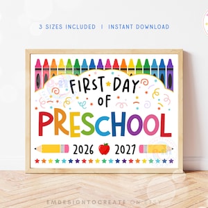 Può includere: Un cartello rettangolare con cornice in legno che riporta la scritta FIRST DAY OF PRESCHOOL in lettere nere e multicolori. Il design include una fila di pastelli colorati in alto, due matite gialle, una mela rossa e una fila di stelle in basso.