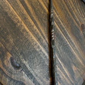 May include: A dark, hand-forged metal wand with a pointed tip. The wand features a twisted design on the lower half and a series of square-like shapes on the upper half. The wand is set against a wooden background.