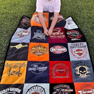 May include: A patchwork quilt made from Harley-Davidson t-shirts. The quilt features various logos and graphics in orange, black, red, and white. The quilt is spread out on green grass.