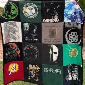May include: A patchwork quilt constructed from various graphic t-shirts featuring superhero and pop culture themes. The quilt displays diverse prints including Arrow, Breaking Benjamin, Black Lightning, and Avengers logos, alongside text like "Who needs swag when you got the heart of the cards" and "I have come home with only one goal to save my city."