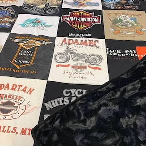 May include: A patchwork quilt constructed from various Harley-Davidson t-shirts featuring motorcycle graphics, logos, and location text such as Sturgis, Cabo San Lucas, and Jacksonville. The quilt has a black, textured, plush fabric backing.