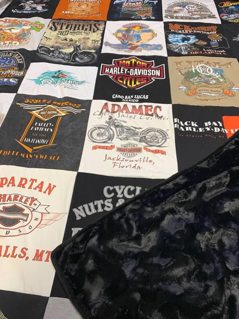 May include: A patchwork quilt constructed from various Harley-Davidson t-shirts featuring motorcycle graphics, logos, and location text such as Sturgis, Cabo San Lucas, and Jacksonville. The quilt has a black, textured, plush fabric backing.