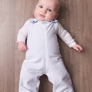 May include: A baby wearing a white jumpsuit with blue trim and a long-sleeved white shirt. The baby is lying on a wooden floor, looking up at the camera.