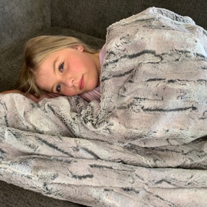 May include: A young person is lying on a gray couch, wrapped in a soft, gray and white faux fur throw blanket. The blanket has a striped pattern and is very fluffy.