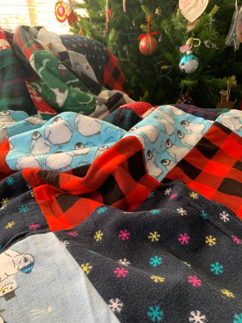 May include: A patchwork quilt featuring various fabric patterns including light blue panels with penguin illustrations, red and black buffalo plaid squares, and a dark navy blue fabric decorated with small, colorful embroidered snowflake motifs.