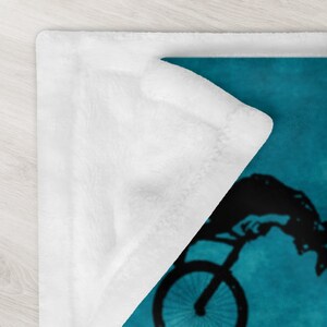 K&ouml;nnte beinhalten: Eine tealblaue Fleece-Decke mit einer schwarzen Silhouette einer Person, die einen Stunt auf einem Fahrrad macht. Die Decke hat ein wei&szlig;es Fleece-Futter.
