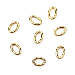 May include: Nine gold oval jump rings, a type of jewellery finding, are scattered on a white background.