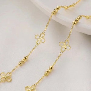 Peut inclure: Collier doré avec un design de chaîne délicat. Le collier présente de petites perles rondes et des pendentifs en forme de trèfle à quatre feuilles. Le bijou est présenté sur une surface blanche.