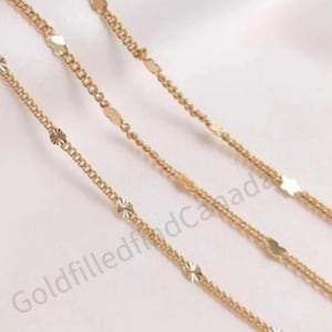 Puede incluir: Tres cadenas de oro con pequeños eslabones decorativos y con patrones. Las cadenas están superpuestas sobre una superficie blanca.