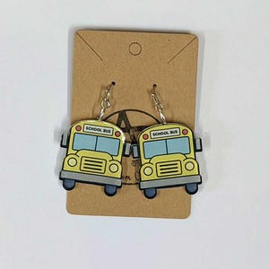 May include: A pair of yellow school bus earrings with black and blue accents. Each earring has the words "SCHOOL BUS" printed on the top. The earrings are hanging on a brown card.