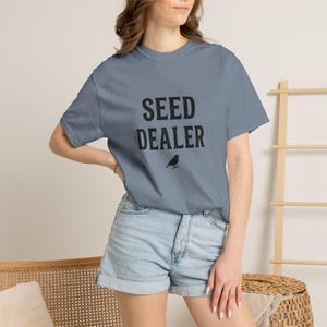 以下が含まれることがあります： 胸元に黒い文字で「SEED DEALER」とプリントされ、その下に鳥の小さな黒いグラフィックが描かれた、スレートブルーの半袖Tシャツ。クルーネックで、標準的なフィット感のTシャツです。