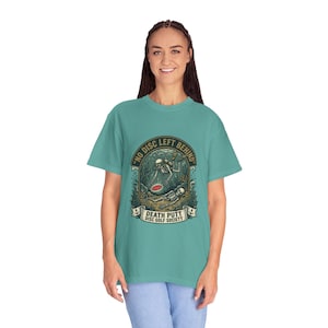 Pode incluir: Uma camiseta verde-azulado com um design gráfico com o texto "NO DISC LEFT BEHIND" e "DEATH PUTT DISC GOLF SOCIETY". O design mostra esqueletos jogando disc golf debaixo d'água. A camiseta é feita de um material macio e confortável.