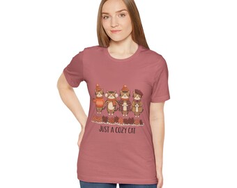 May include: A mauve t-shirt featuring a graphic of four cartoon cats wearing fall attire, standing in front of autumn leaves. The text "JUST A COZY CAT" is printed below the cats.