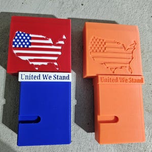 May include: Two 3D printed boxes with the American flag and the words "United We Stand" on the front. The boxes are red, white, and blue on the left and orange on the right.