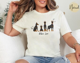 May include: Cream-colored t-shirt featuring a graphic of four Doberman Pinschers in various poses, with hearts and the text "Dobie Love" in black. The shirt is made of a soft material.