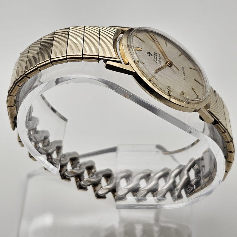 May include: A gold-toned Zodiac automatic wristwatch with a silver-colored face and a gold-toned expanding band. The watch is displayed on a clear, curved stand, showcasing its design and details. The watch face has the Zodiac logo and the words "automatic" and "25 jewels".