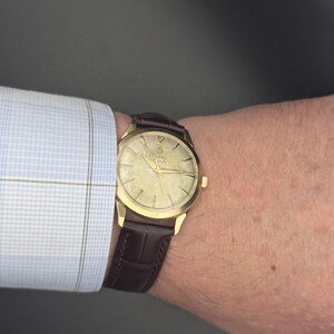 Pode incluir: Um rel&oacute;gio Zodiac dourado com uma pulseira de couro castanho. O mostrador do rel&oacute;gio tem ponteiros e marcadores dourados, com o n&uacute;mero 12 no topo. O rel&oacute;gio est&aacute; num pulso, ao lado de uma camisa xadrez azul clara.
