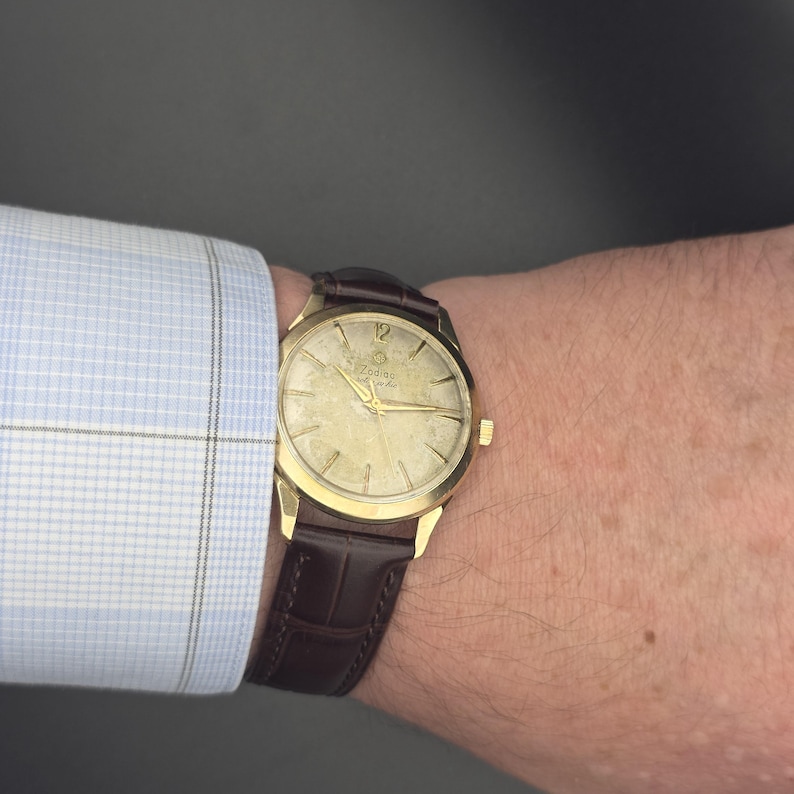 Pode incluir: Um rel&oacute;gio Zodiac dourado com uma pulseira de couro castanho. O mostrador do rel&oacute;gio tem ponteiros e marcadores dourados, com o n&uacute;mero 12 no topo. O rel&oacute;gio est&aacute; num pulso, ao lado de uma camisa xadrez azul clara.