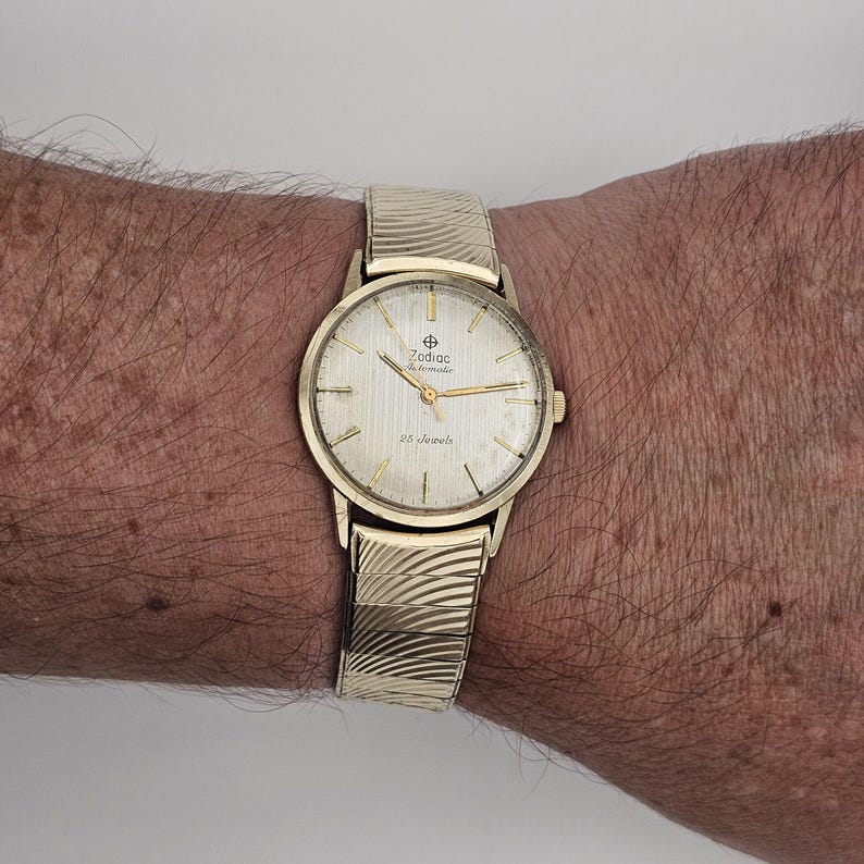 May include: A gold-toned Zodiac automatic wristwatch with a textured band. The watch face is light-colored with gold hands and markers. The watch face has the text "Zodiac Automatic" and "25 jewels".