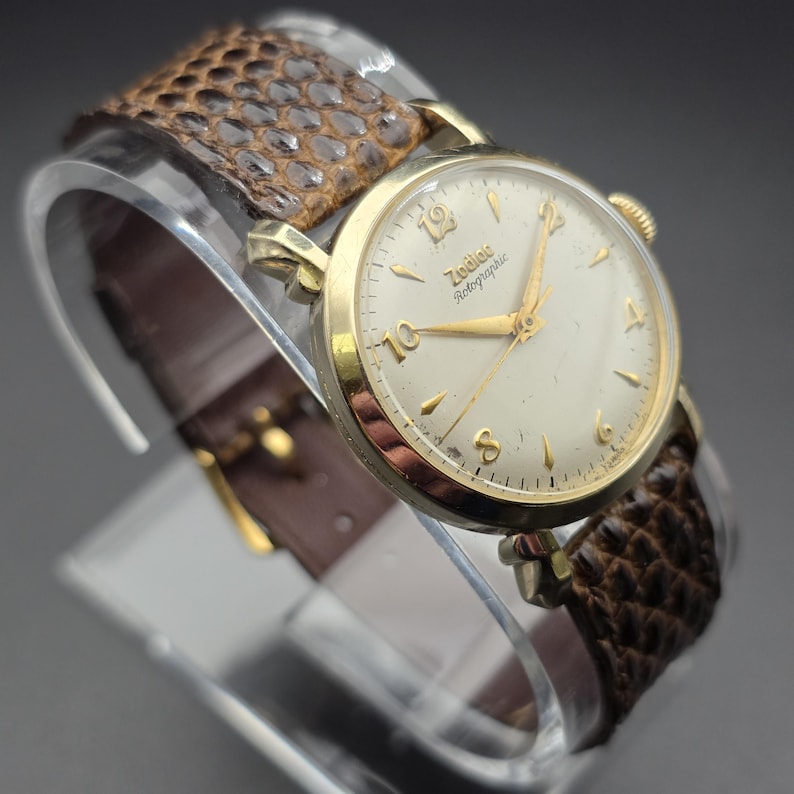 K&ouml;nnte beinhalten: Eine Vintage Zodiac Rotographic Armbanduhr mit goldfarbenem Geh&auml;use und cremefarbenem Zifferblatt. Die Uhr hat goldene Zeiger und Stundenmarkierungen. Sie hat ein braunes Lederarmband mit Schlangenmuster.